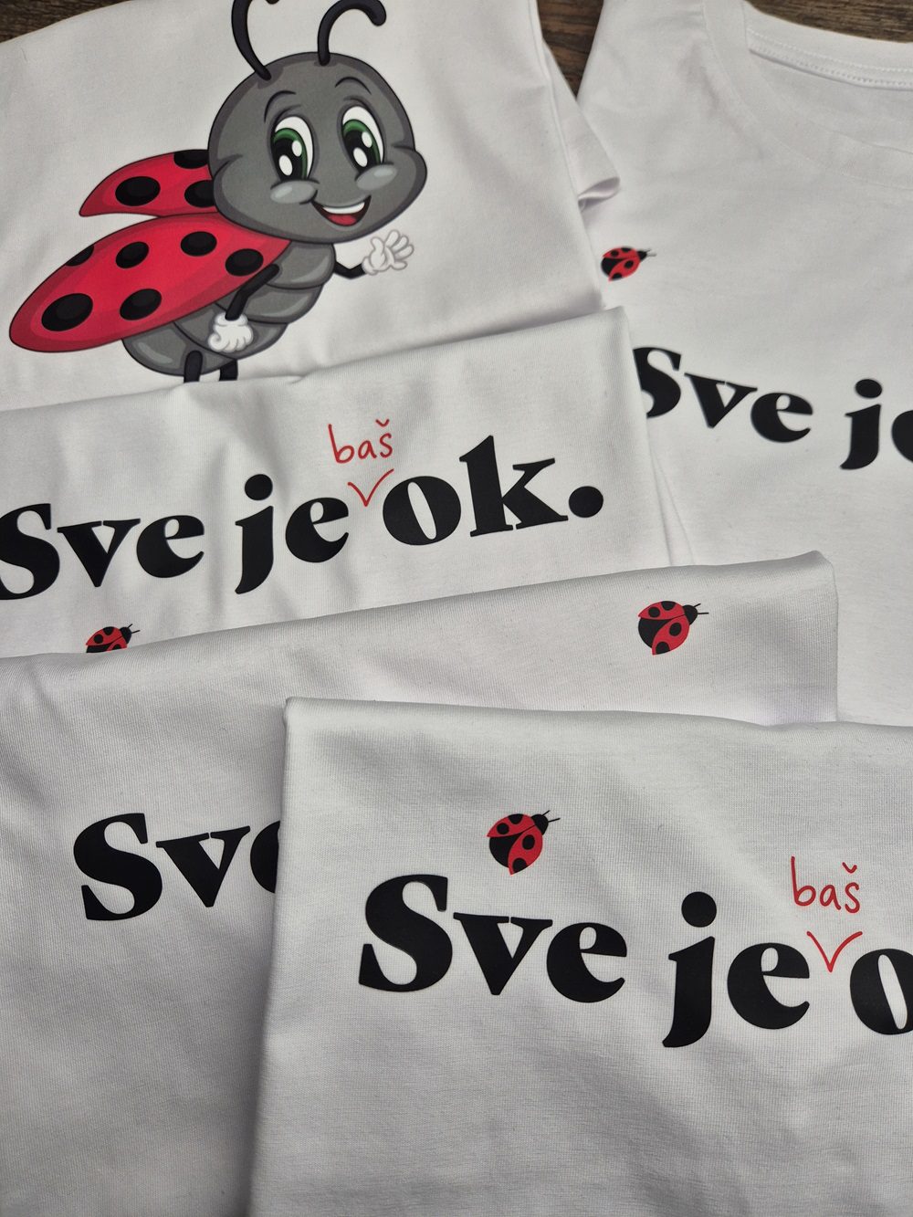 Bijele majice s tiskom natpisa "Sve je baš OK" i motivom bubamare raspoređene na drvenoj površini. Jedna majica sadrži ilustraciju simpatične bubamare s osmijehom i podignutom rukom. Dizajn kombinira crnu i crvenu boju, stvarajući upečatljiv vizualni identitet.